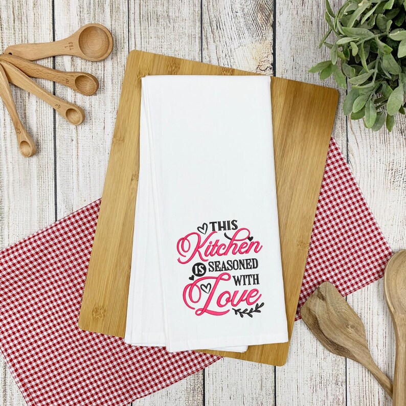 May include: White kitchen towel with the text "This Kitchen is Seasoned with Love" in black and pink lettering with a heart design.
