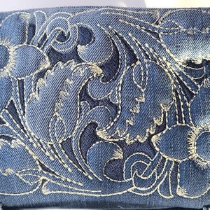 Könnte beinhalten: Nahaufnahme eines Jeansstoffs mit aufwendiger weißer Blumenstickerei. Das Design zeigt detaillierte Blumen und wirbelnde Muster, die eine strukturierte und dekorative Oberfläche schaffen. Der Jeansstoff ist mittelblau.