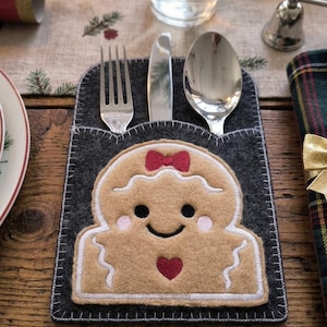 May include: A dark gray felt cutlery holder featuring an embroidered tan gingerbread person with a red bow, pink cheeks, and a red heart. The holder contains a silver fork, a silver knife, and a silver spoon tucked into the top pocket.