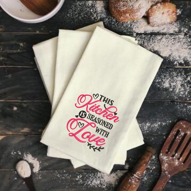 May include: Set of four white kitchen towels with pink and black embroidery that reads "This Kitchen is Seasoned with Love".