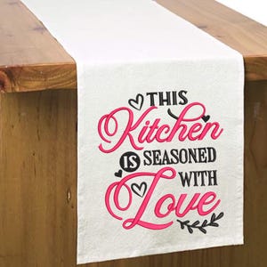 May include: White table runner with pink and black embroidery that says "This Kitchen is Seasoned with Love".