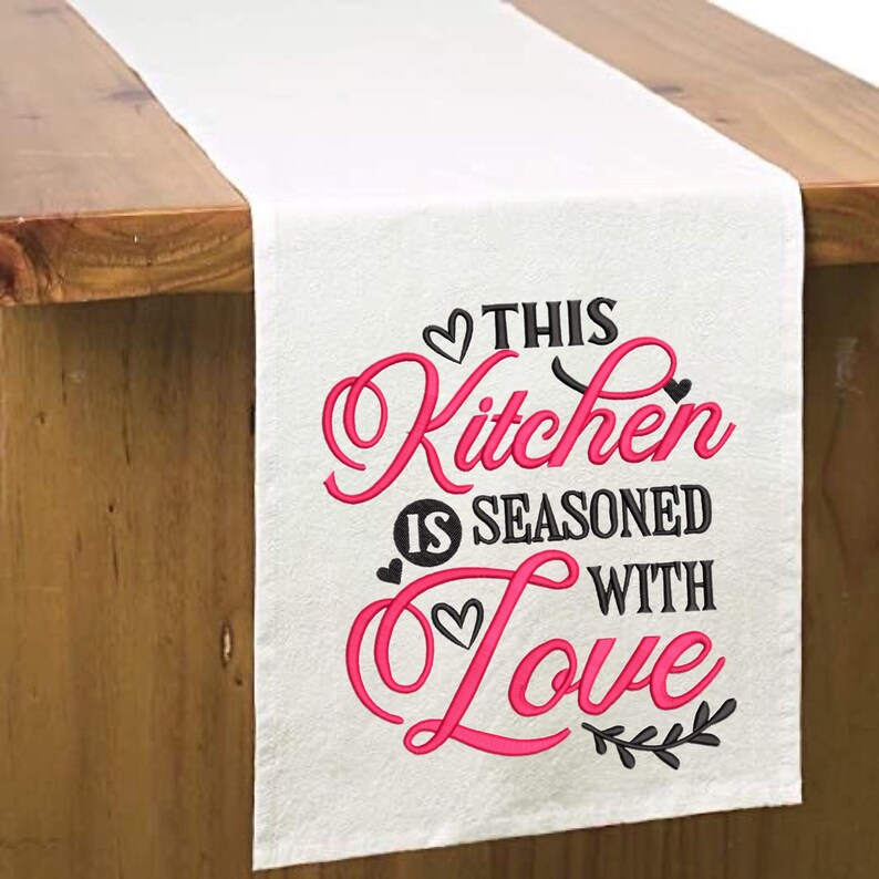 May include: White table runner with pink and black embroidery that says "This Kitchen is Seasoned with Love".