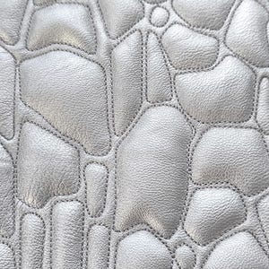 Pode incluir: Close-up de um material de couro prateado com um padrão acolchoado. O design apresenta formas irregulares e arredondadas costuradas com fio cinza, criando uma superfície texturizada. O couro tem um brilho sutil.