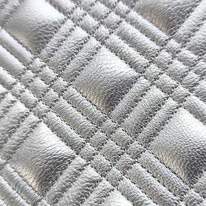 May include: Close-up of a silver quilted fabric with a diamond pattern. The fabric has a textured, metallic appearance with visible stitching creating the quilted effect. The pattern is consistent across the surface.