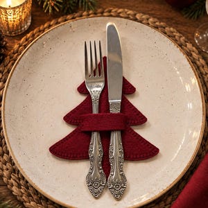 May include: A cream-colored ceramic plate with dark speckles holds a dark red felt Christmas tree cutout. A silver fork and a silver knife with ornate floral handles are tucked into the center of the felt tree, resting vertically on the plate.