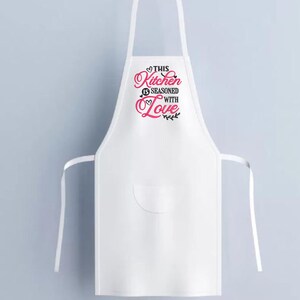 May include: White kitchen apron with a pink heart and black text that reads "This Kitchen is Seasoned with Love".