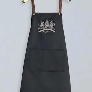 May include: Black apron with brown straps and a large pocket. The apron has a white design of three Christmas trees with the text "Merry Christmas" underneath.