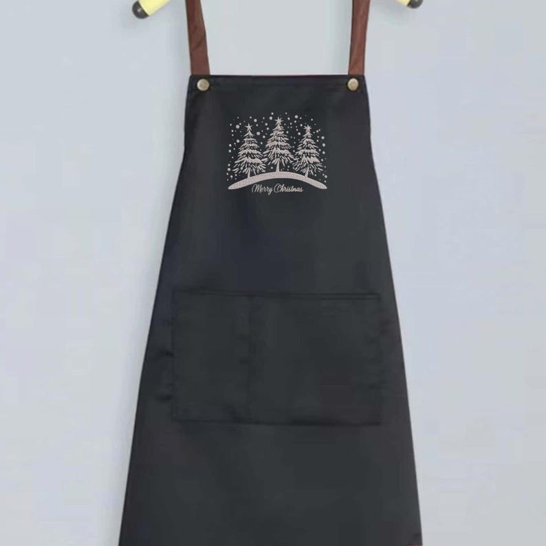 May include: Black apron with brown straps and a large pocket. The apron has a white design of three Christmas trees with the text "Merry Christmas" underneath.