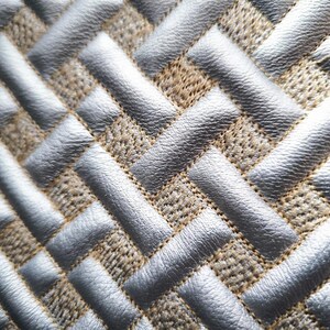 Puede incluir: Una tela texturizada con un patrón geométrico repetitivo de tiras plateadas metálicas en relieve tejidas sobre un material base de color tostado y grano grueso. El diseño crea un efecto de tejido de cesta con líneas diagonales.