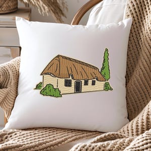 May include: Embroidered cushion featuring a white cottage with a thatched roof, brown door, and two windows. The cottage is surrounded by green bushes and a tall, green tree.