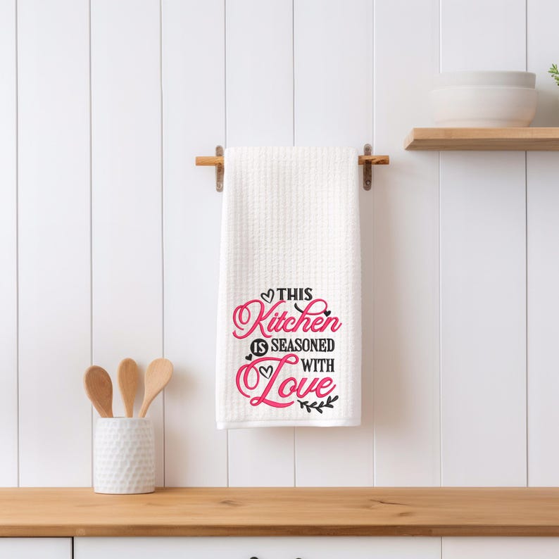 May include: White kitchen towel with the text "This Kitchen is Seasoned with Love" in black and pink lettering with a small heart design and a sprig of leaves.