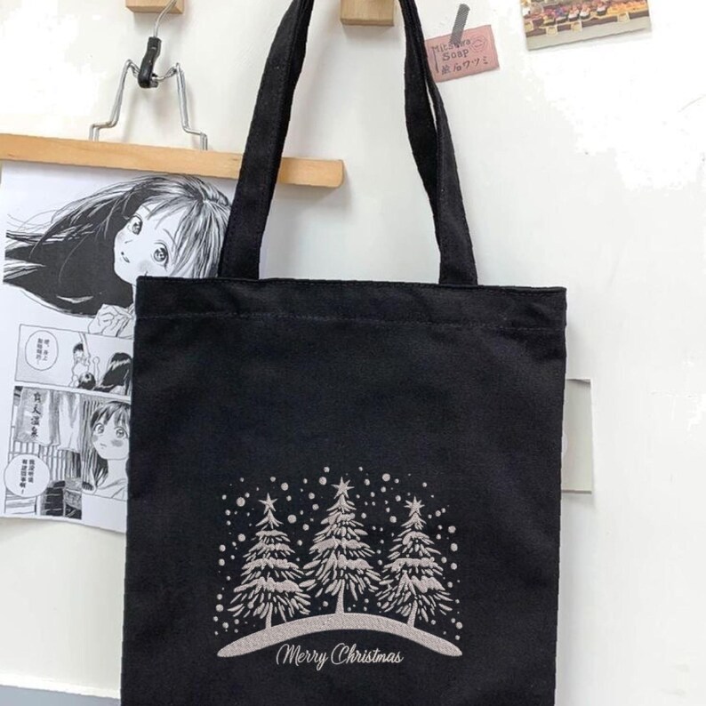 May include: Black canvas tote bag with a white design of three Christmas trees and snowflakes. The text "Merry Christmas" is printed below the trees.