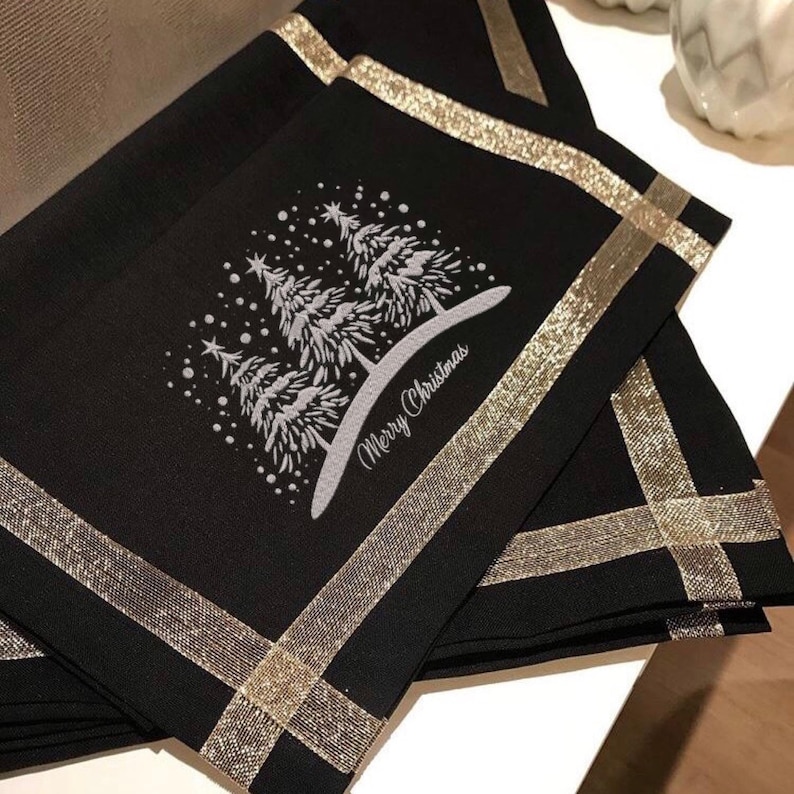 May include: Black fabric placemats with gold glitter trim and a white embroidered design of Christmas trees and the text "Merry Christmas".