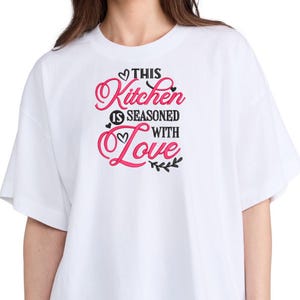 May include: White t-shirt with the text "This Kitchen is Seasoned with Love" in black and pink lettering.