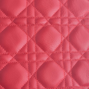 Puede incluir: Primer plano de una textura de cuero acolchado rojo. El patrón presenta un diseño repetitivo de formas de diamante y cuadrados redondeados, todos cosidos con hilo rojo a juego. El cuero tiene una superficie ligeramente texturizada.