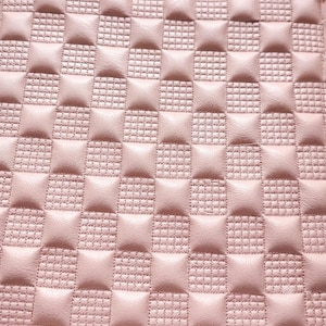 Puede incluir: Un material de color rosa claro con un patrón de cuadros repetitivo. El diseño consiste en secciones cuadradas lisas y elevadas que se alternan con secciones cuadradas hundidas con una textura fina en forma de rejilla en toda la pieza.