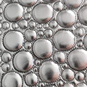 Może przedstawiać: Zbliżenie srebrnej, teksturowanej powierzchni z wzorem wypukłych, okrągłych kształtów. Okręgi różnią się wielkością i są obrysowane ciemniejszą, szytą obwódką. Całkowity efekt to nowoczesny, metaliczny design.