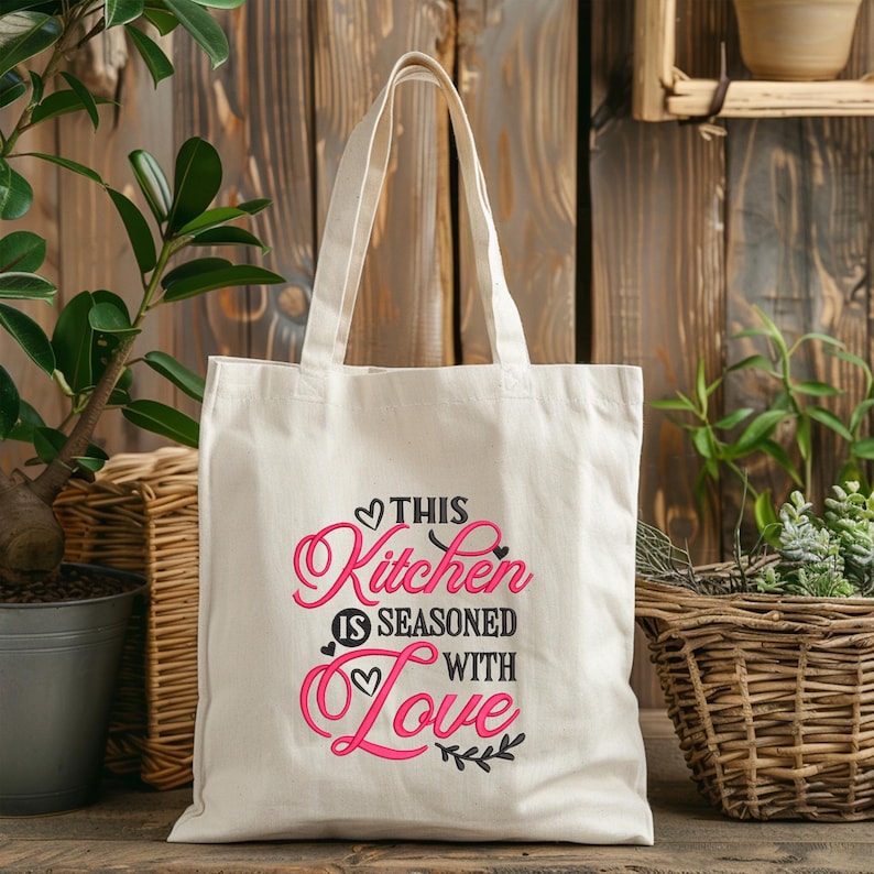 May include: A white canvas tote bag with black and pink text that reads "This Kitchen is Seasoned with Love". The bag has black handles and is sitting on a wooden surface.