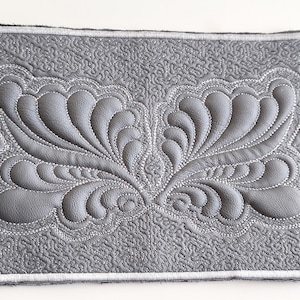 May include: A rectangular, gray, quilted placemat with a central, symmetrical design. The design features a raised, stylized feather or leaf pattern, stitched with white thread. The border is also stitched with white thread.