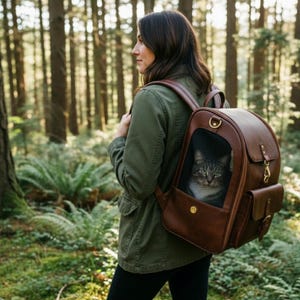 May include: A brown leather cat backpack with a mesh window, worn by a person in a forest. The backpack has a gold-colored clasp and a small side pocket. A gray tabby cat is visible inside the backpack.