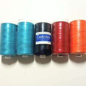 May include: Five cylindrical spools of sewing thread arranged in a row. The colors from left to right are two shades of teal, one black spool with a white label reading Essential from ConnectingThreads.com, one red, and one orange spool of thread.