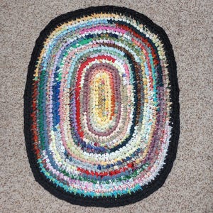 May include: Oval-shaped, handmade rag rug with a black border. The interior features concentric rings of colorful fabric strips in various patterns and hues, including red, blue, yellow, and green. A unique, decorative floor covering.