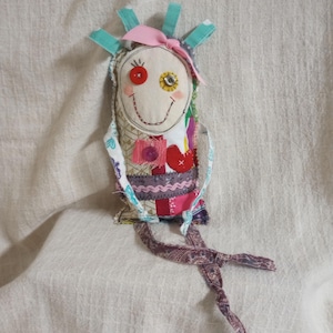 May include: A handmade fabric doll featuring a cream-colored face with a red button eye, a yellow button eye, and a stitched smile. The body is constructed from various patterned fabric patches in shades of pink, red, and purple, accented with teal fabric strips at the top and a long, knotted paisley-patterned fabric tail at the base.