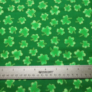 May include: A green fabric featuring a repeating pattern of light green shamrocks with gold glitter outlines. A metal Westcott ruler is positioned across the bottom, showing measurements in inches and centimeters for scale reference.