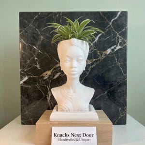 May include: A white bust planter in the shape of a woman's head, holding a green plant. The planter sits on a wooden base in front of a black and gray marble slab. A business card reads "Knacks Next Door - Handcrafted & Unique".