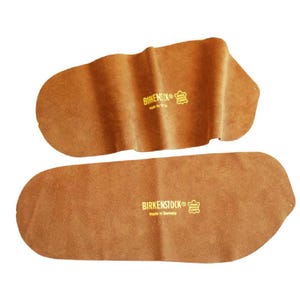 May include: Two brown leather Birkenstock insoles. The insoles are a light brown color and have the Birkenstock logo printed in gold. The insoles are designed to fit the shape of a foot.