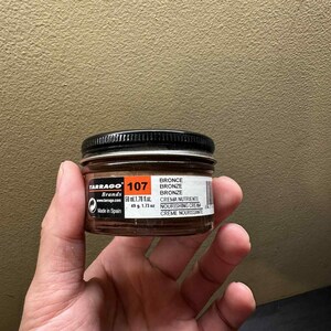 May include: A small glass jar of Tarrago Brand shoe cream in the color "Bronze." The jar has a black lid and a white label with product information, including "107" and "50 ml / 1.78 fl. oz."