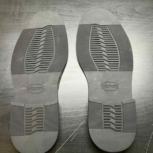 May include: Two black shoe soles with a textured tread pattern. The soles are made of a durable material and feature the "Vibram" logo. The soles are designed for footwear and provide grip and traction.