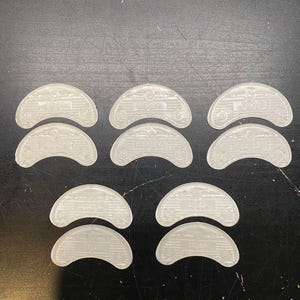 May include: A collection of clear, semi-circular plastic pieces, each with a curved top and a flat bottom. The pieces have a textured surface with a series of small, circular and rectangular details. The objects are arranged on a dark surface.