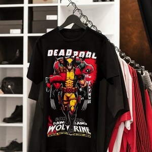 Puede incluir: Camiseta negra con un gr&aacute;fico de Wolverine cargando a Deadpool. El dise&ntilde;o incluye las palabras "DEADPOOL" y "WOLVERINE" en blanco y rojo, con texto adicional en la parte inferior. La camiseta est&aacute; colgada en una percha con otras prendas.