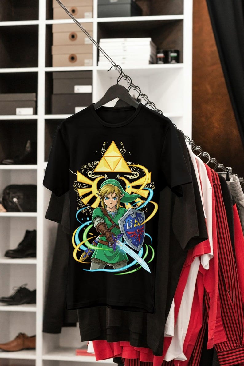 May include: Black t-shirt featuring a colorful graphic of a video game character with a sword and shield. The design includes a golden triangle and swirling blue and yellow accents. The shirt is hanging on a black hanger.