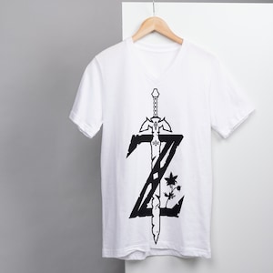 May include: White V-neck t-shirt with a black graphic of a sword and the letter "Z" on the front.