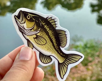 May include: A sticker featuring a detailed illustration of a largemouth bass fish. The fish is depicted in shades of yellow, black, and white, with intricate scale and fin details. The sticker is held against a blurred background of water and greenery.