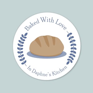 May include: A round sticker with a light blue background. The sticker has a white circle with a loaf of bread in the center. The text "Baked With Love" arches above the bread, and "In Daphne's Kitchen" is below. Blue leaf sprigs frame the bread.