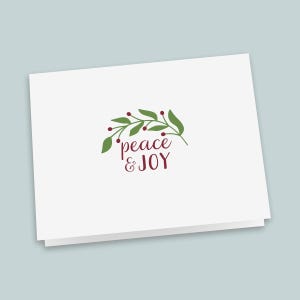 May include: A white folded greeting card featuring a centered graphic of a green leafy branch with small red berries. Below the branch, the text "peace & JOY" is printed in a dark red script and sans-serif font combination.