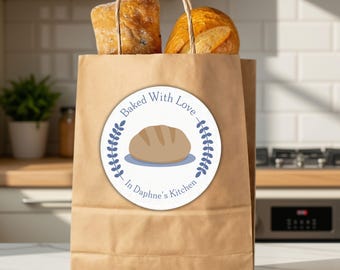 May include: A brown paper bag with twisted paper handles containing two loaves of bread. A circular white sticker on the front features a blue illustration of a loaf of bread, blue leaf accents, and the text Baked With Love In Daphne's Kitchen.