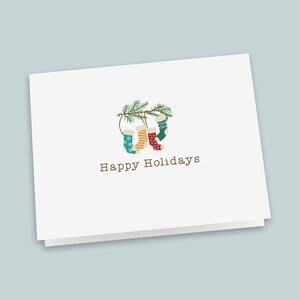 May include: A white greeting card featuring a central illustration of four colorful Christmas stockings hanging from a green pine branch. The stockings are teal, yellow, red, and green with various patterns. The text Happy Holidays is printed below.