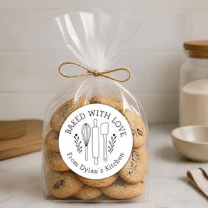 May include: A clear plastic bag filled with round, light brown cookies, tied at the top with a tan twine bow. A circular white sticker on the front features black line art of a whisk, rolling pin, and spatula, with the text BAKED WITH LOVE From Dylan's Kitchen.