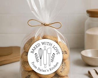 May include: A clear plastic bag filled with round, light brown cookies, tied at the top with a tan twine bow. A circular white sticker on the front features black line art of a whisk, rolling pin, and spatula, with the text BAKED WITH LOVE From Dylan's Kitchen.