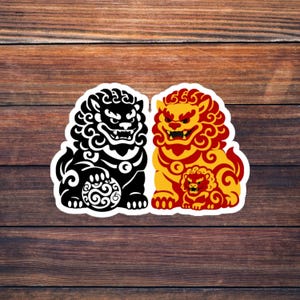 May include: A sticker featuring two guardian lion designs. One lion is black, the other is yellow and red. The lions are stylized with intricate details and sit side-by-side on a wooden background.