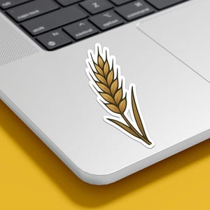 May include: A sticker featuring a detailed illustration of a wheat stalk. The wheat is depicted in shades of brown and gold, with a dark outline. The sticker is placed on a white laptop, with a yellow background.