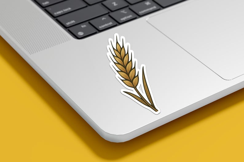May include: A sticker featuring a detailed illustration of a wheat stalk. The wheat is depicted in shades of brown and gold, with a dark outline. The sticker is placed on a white laptop, with a yellow background.