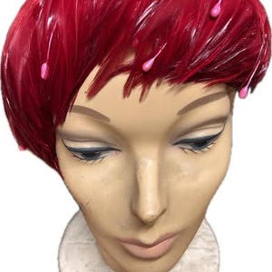 May include: A mannequin head with a red feather hat and pink accents. The hat is made of many small feathers. The mannequin has a neutral expression and is mounted on a white circular base.