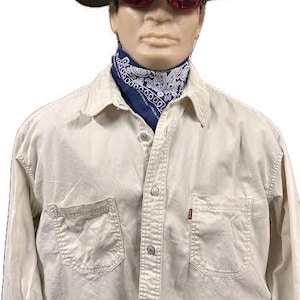 Puede incluir: Maniquí con camisa abotonada de color beige claro con dos bolsillos delanteros y una pequeña etiqueta roja. El maniquí también lleva un sombrero gris, gafas de sol rojas y un pañuelo azul con estampado de cachemira.