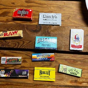 May include: Assortment of rolling papers in various colors and designs, including brands like RAW, Bugler, Zig Zag, and Rollit.  The papers are displayed on a wooden surface.