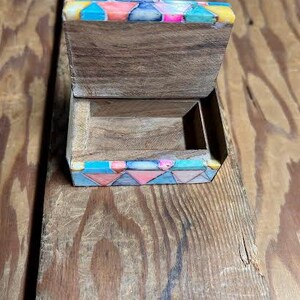 May include: Open wooden box with a hinged lid. The lid and base are decorated with colorful geometric patterns in shades of blue, pink, and yellow. The box is made of wood with a natural grain.